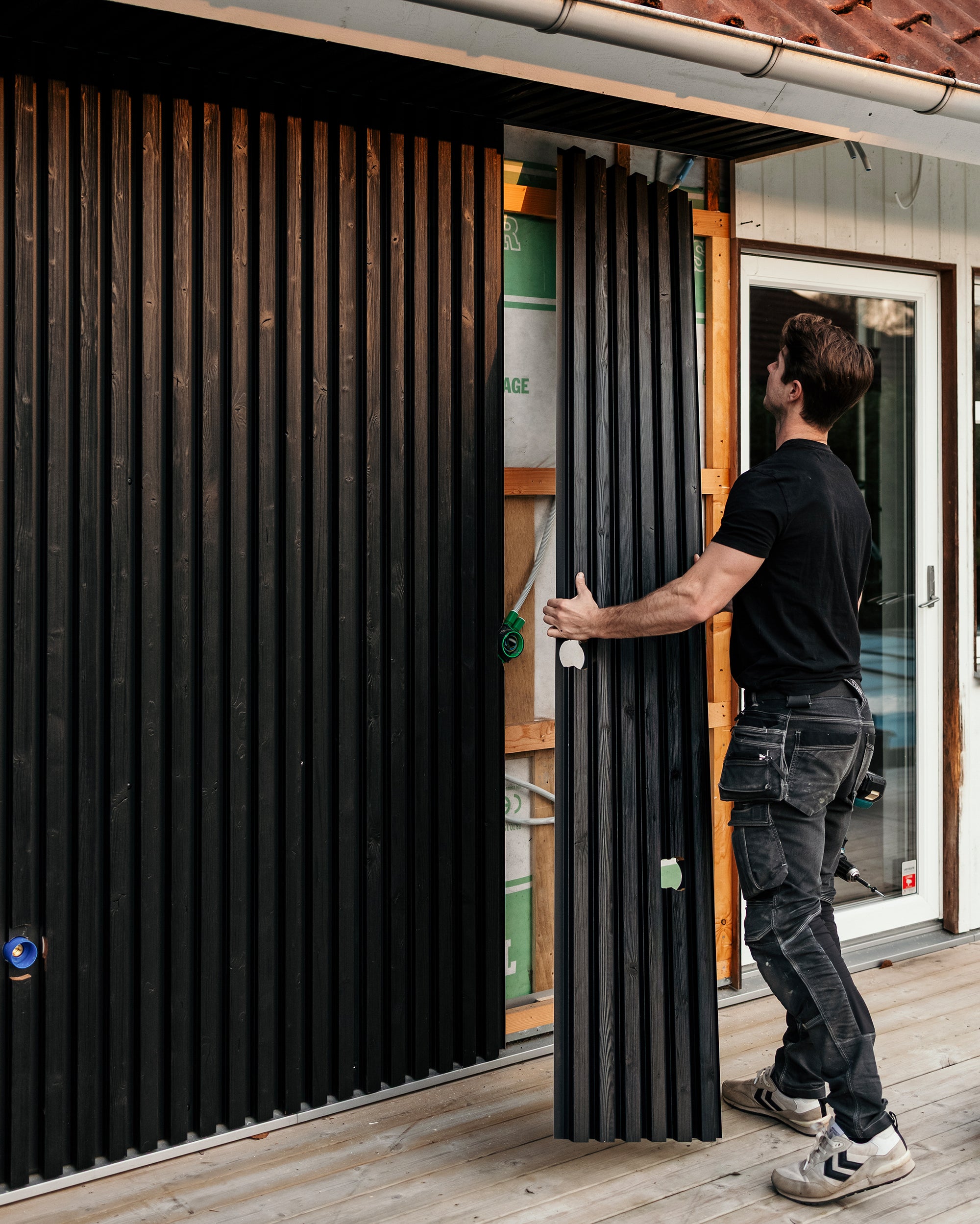 Un uomo installa pannelli per facciata Aluwood neri in posizione verticale sulla parete esterna di una casa mentre è in piedi su un terrazzo di nuova costruzione.